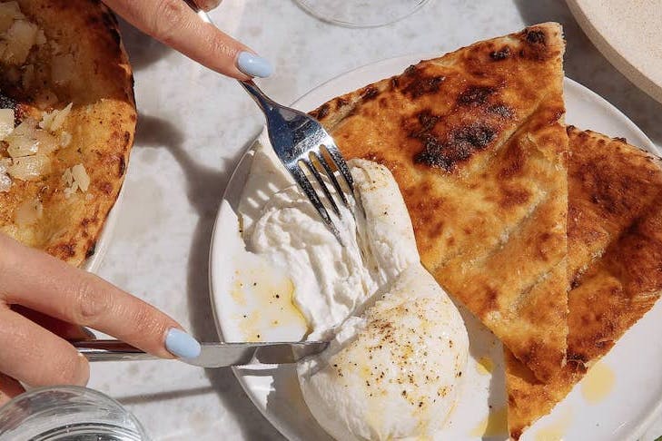 A ball of burrata on a plate with flatbread is pulled apart with cutlery.